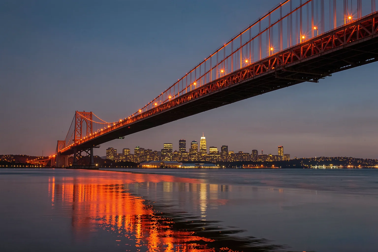 Bay Bridge at Night. An illuminated suspension bridge crosses above a city skyline at night, with orange lights reflected on dark water.