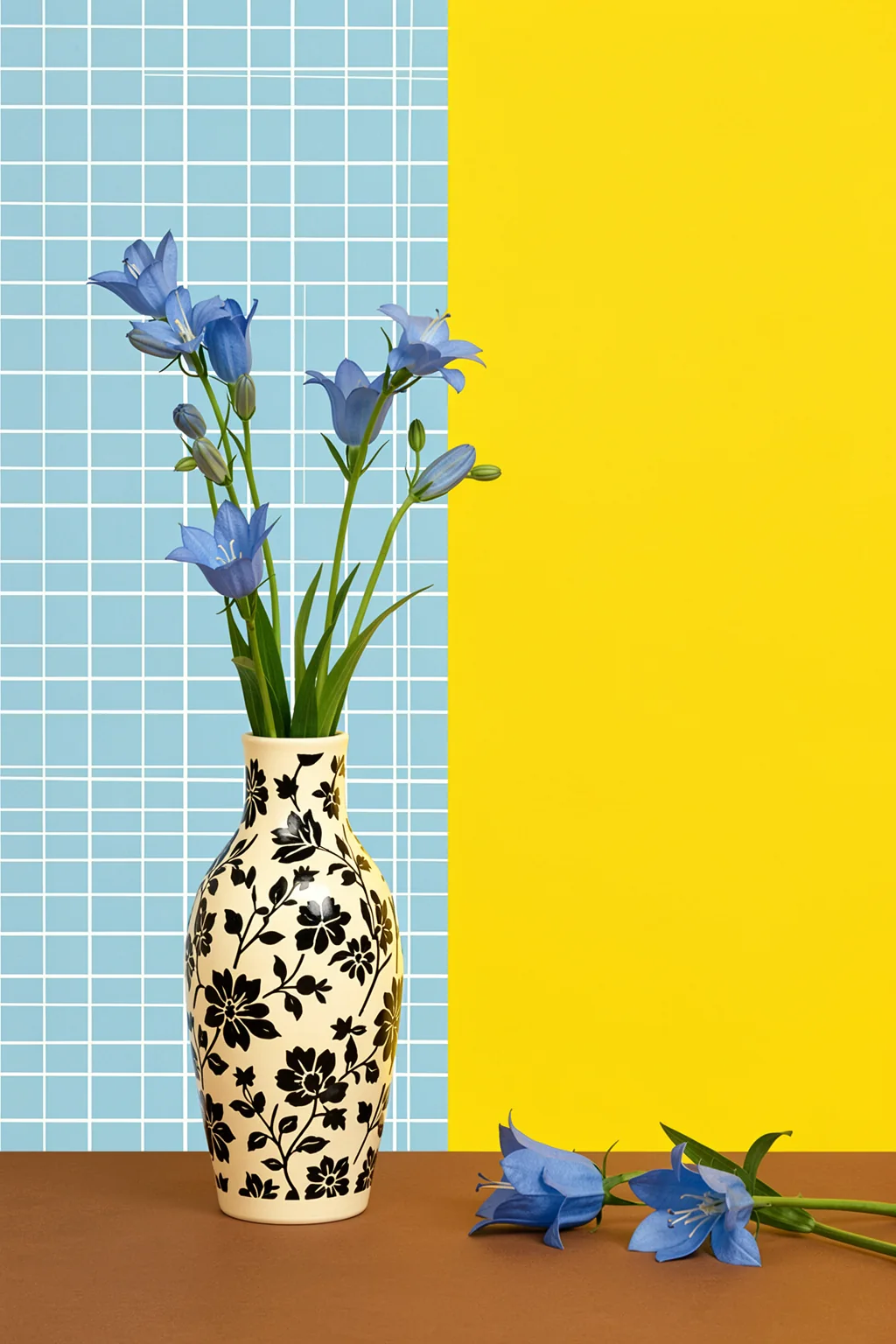 Blue Bellflowers Vase. Blue bellflowers and green stems rise from a patterned cream vase, with a blue tile grid and yellow wall behind.