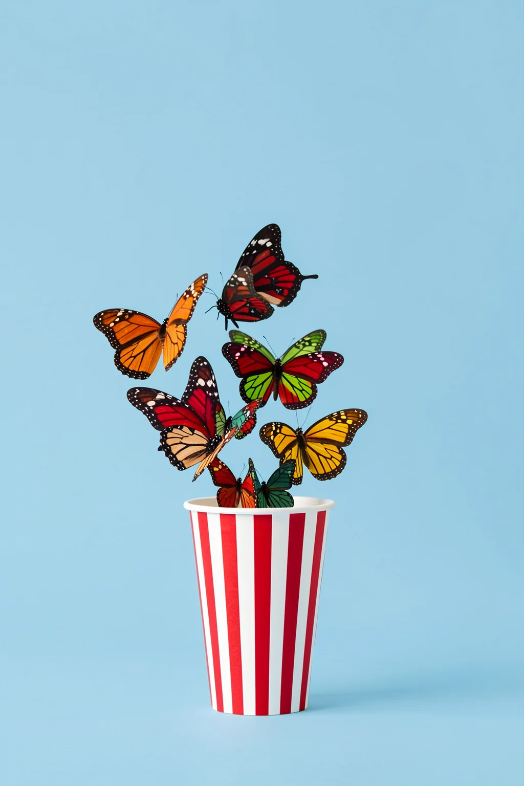 Whimsical Butterfly Pop. Brightly colored butterflies rise from a red-and-white striped cup against a pale blue background.