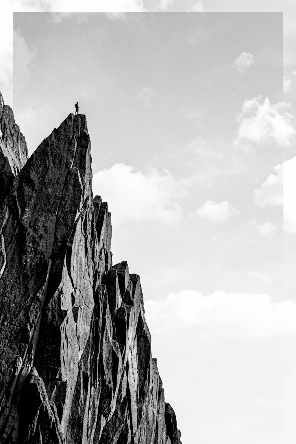 Dramatic Peak Climber. A monochrome photograph shows a steep, jagged rock face rising beneath a bright cloudy sky.