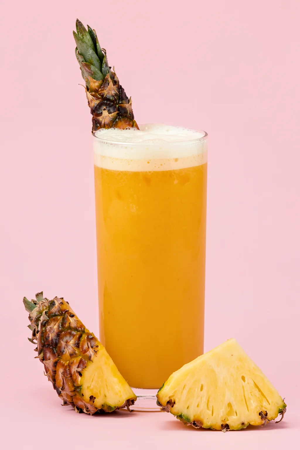 Puerto Rico Piña Colada. An orange-yellow drink in a tall glass is garnished with pineapple against a pink background, with cut pineapple nearby.