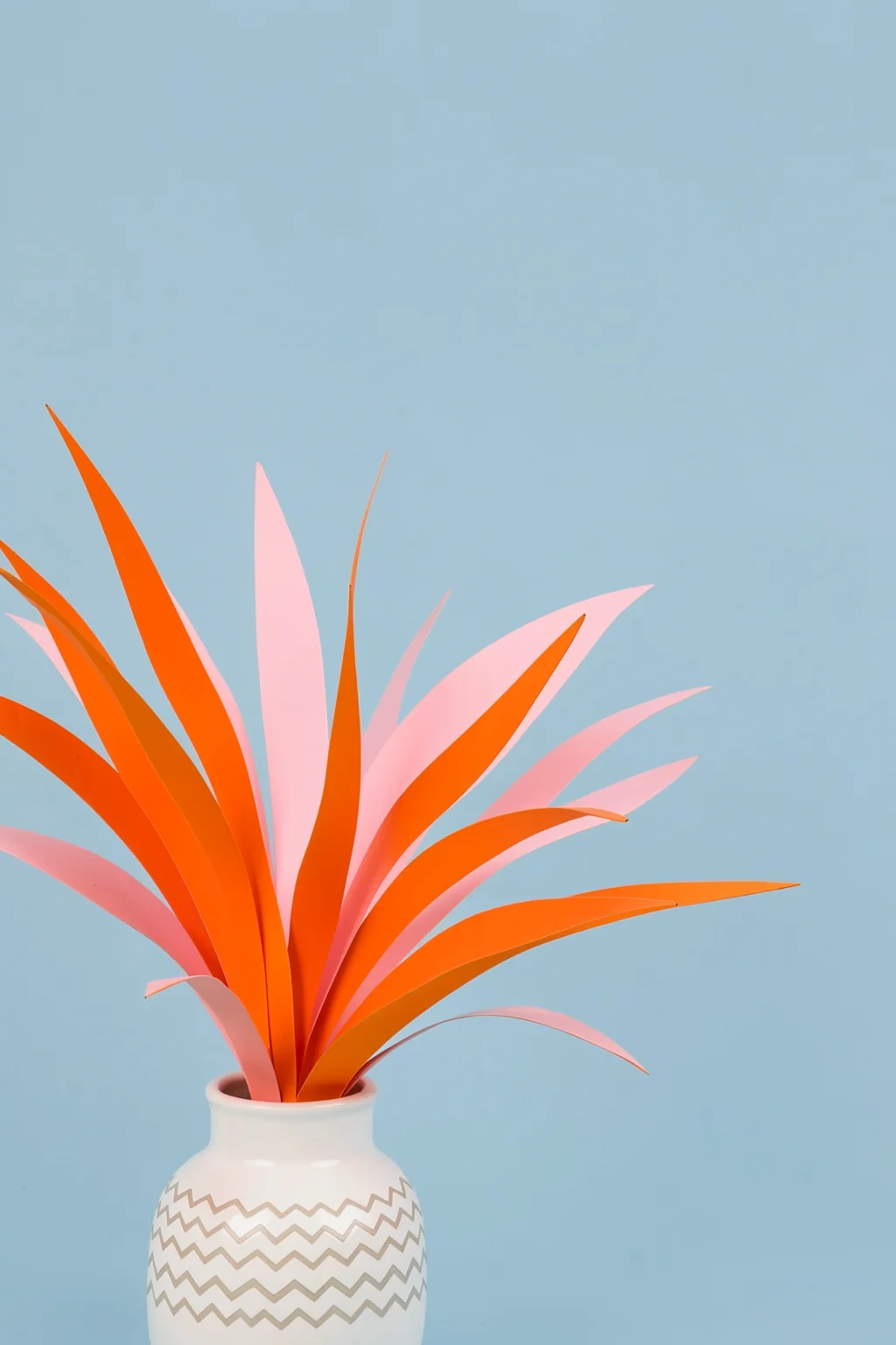Minimal Botanical Still Life. Long orange and pink leaves fan from a white patterned vase against a pale blue background.