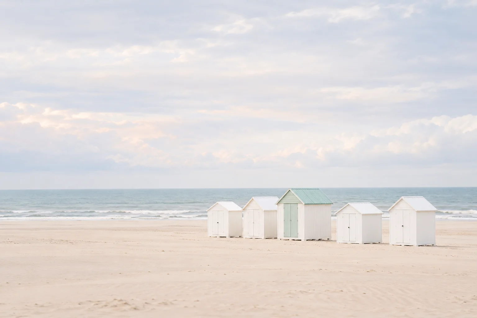 Row of white and pale green beach huts on a wide sand beach under a soft grey sky.