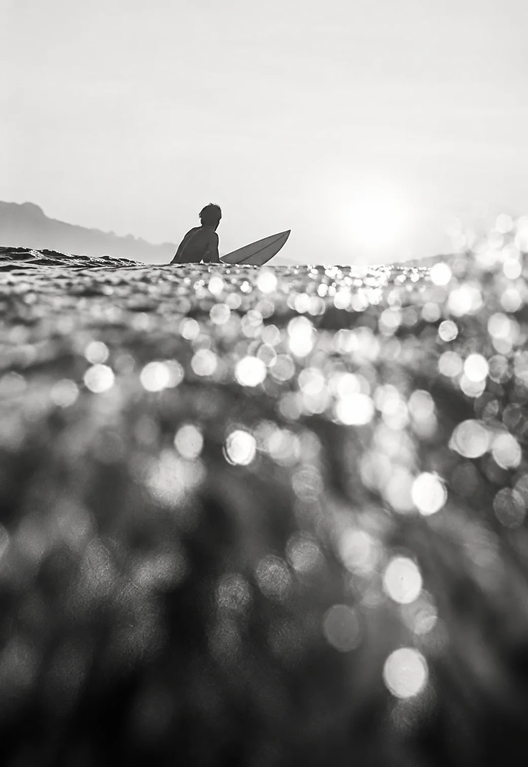 Black-and-white photo of a surfer silhouetted against bright sun over a field of sparkling water droplets.