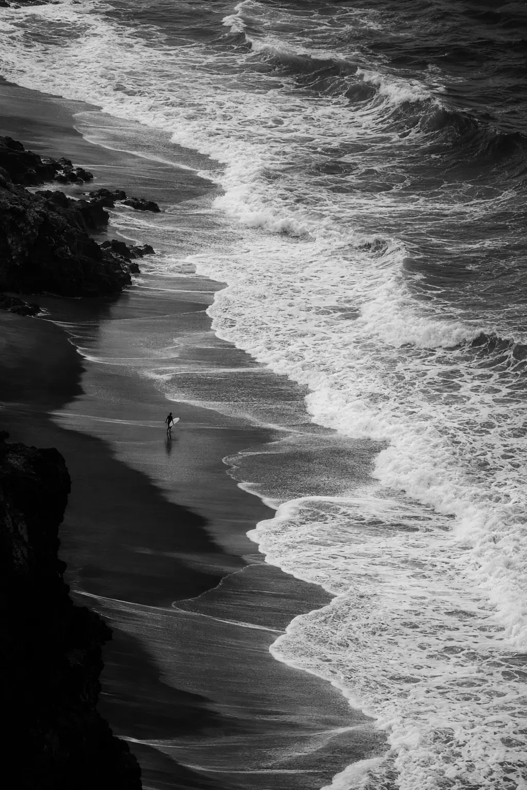 Black-and-white aerial shoreline with rocky edges, churning surf and a tiny figure on the sand.