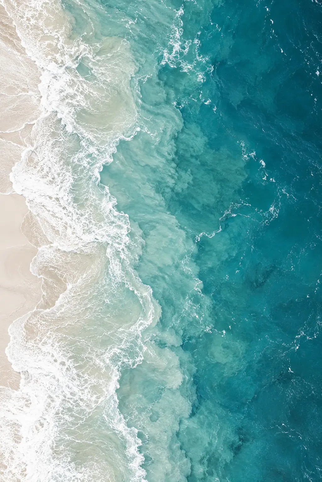 Aerial view of turquoise ocean water meeting pale sand, with white foam tracing the coastline.