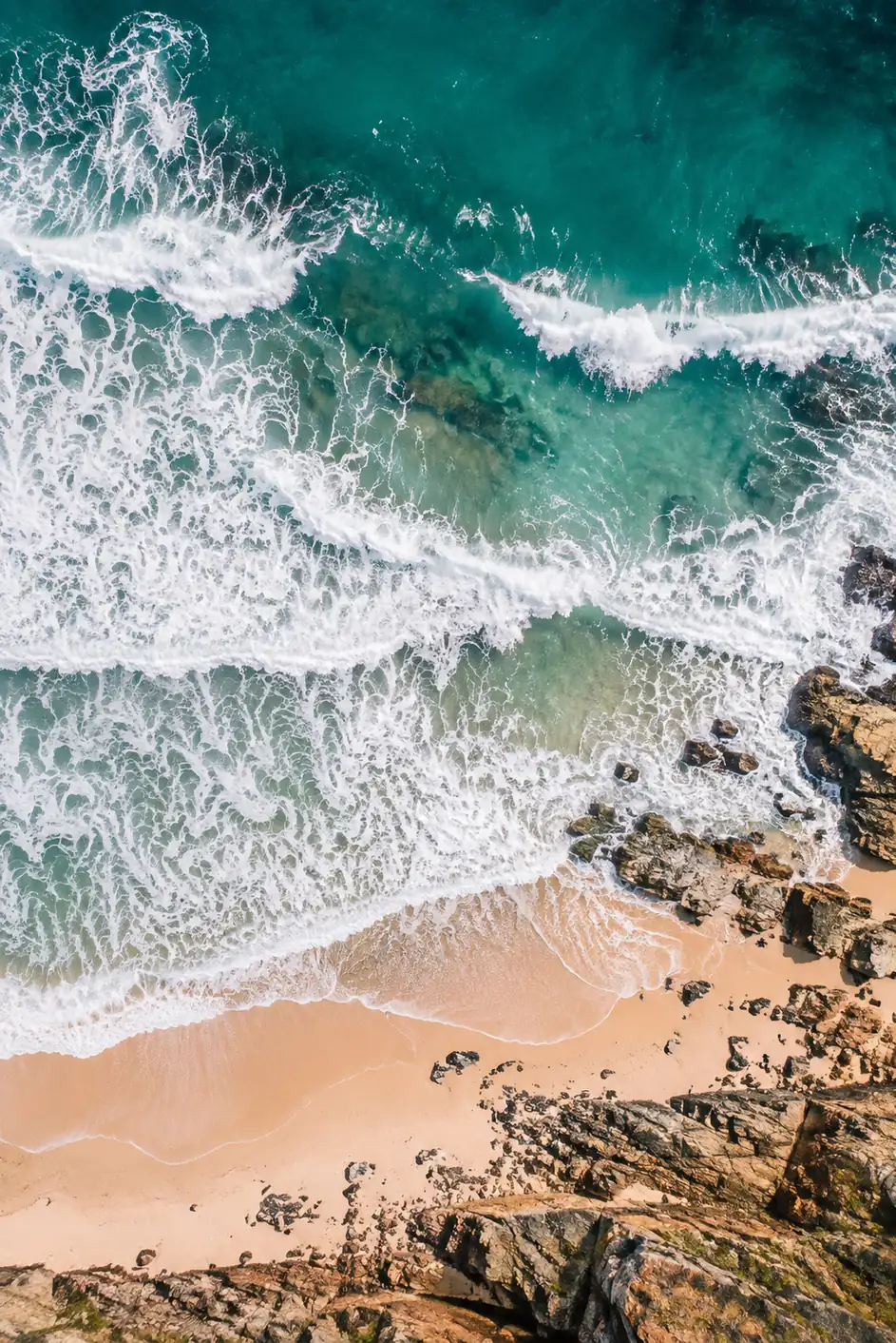 Aerial view of turquoise surf breaking over rocks, with sand and rock textures in the lower corner.