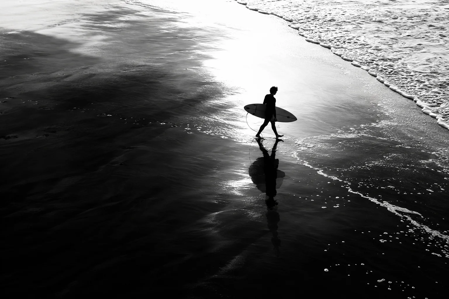 Black-and-white photo of a surfer standing on wet sand with a board, lit from behind by a soft sun.