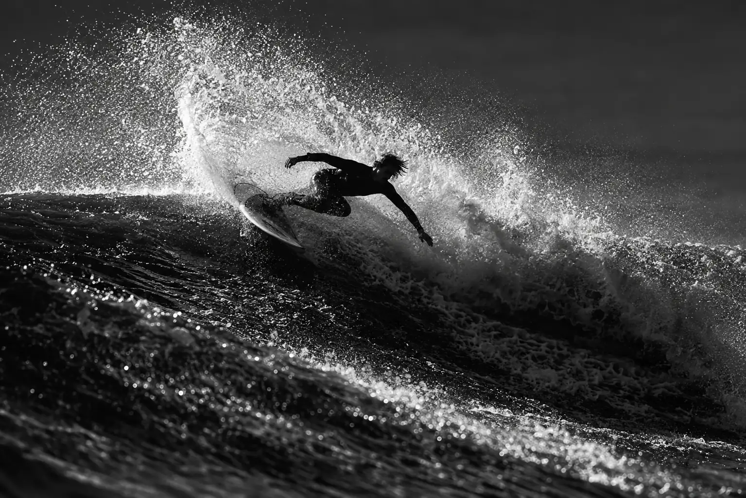 Black-and-white photo of a surfer turning off a wave, with high spray and sharp light across the surface.