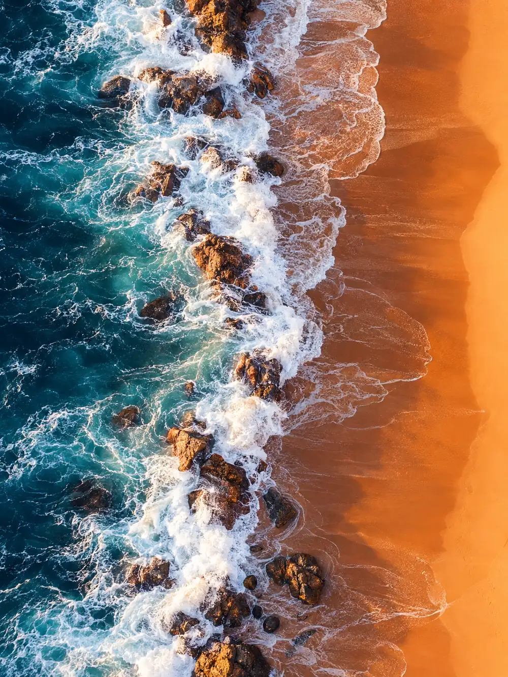 Aerial view of dark rocks and turquoise water meeting a band of warm orange sand.