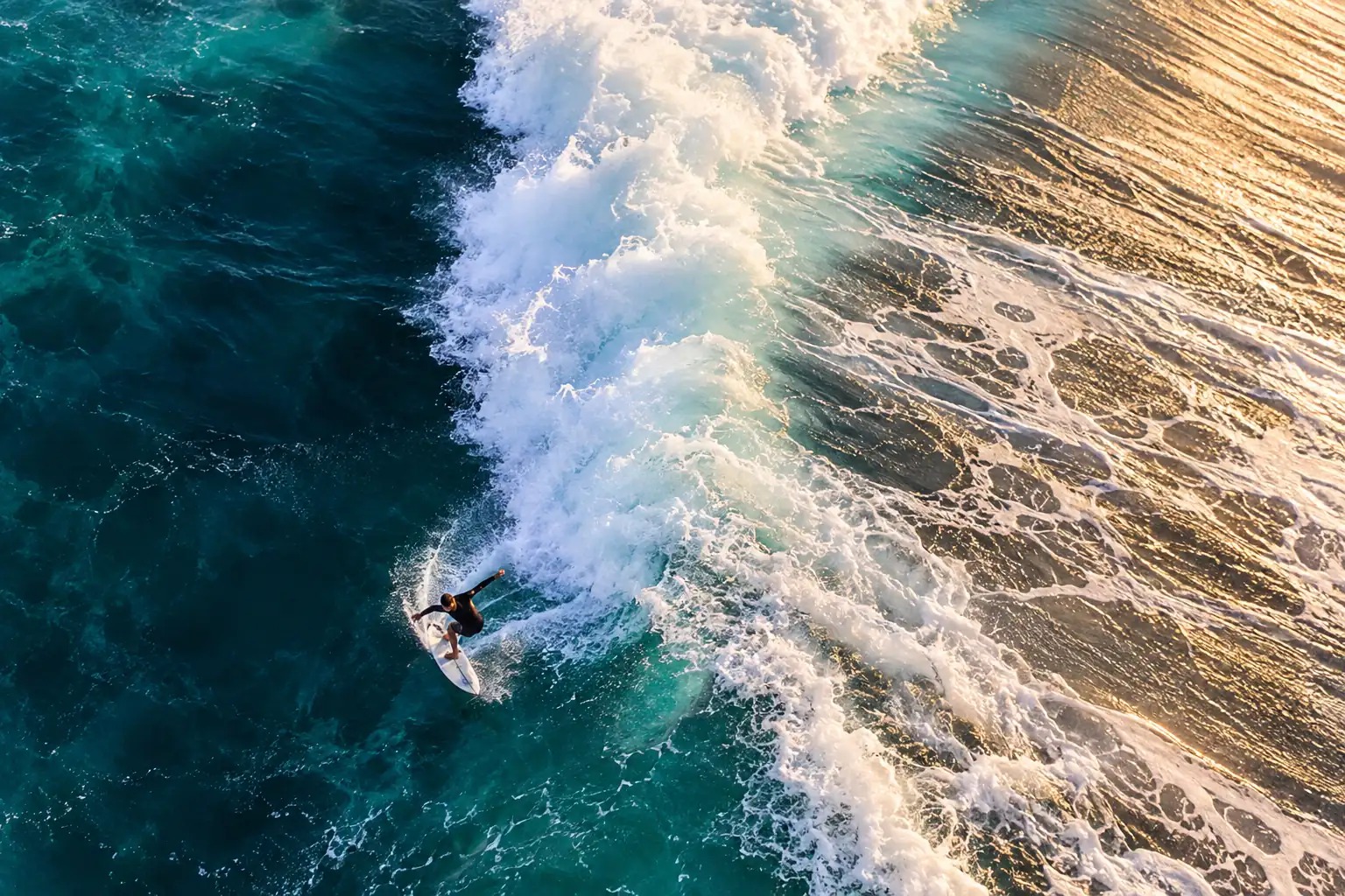 Aerial view of a surfer on a turquoise wave edge with white foam and warm sand below.