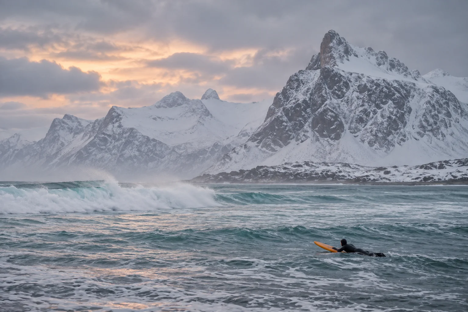 Snow-covered mountains rising above a grey sea, with a surfer paddling in the foreground.