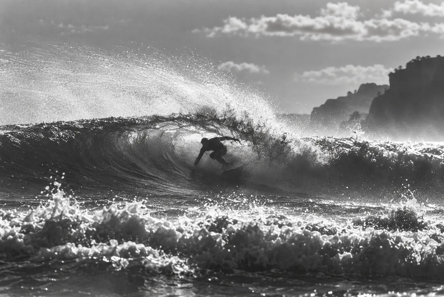 Black-and-white photo of a breaking wave beneath cliffs, with a surfer riding the curl.