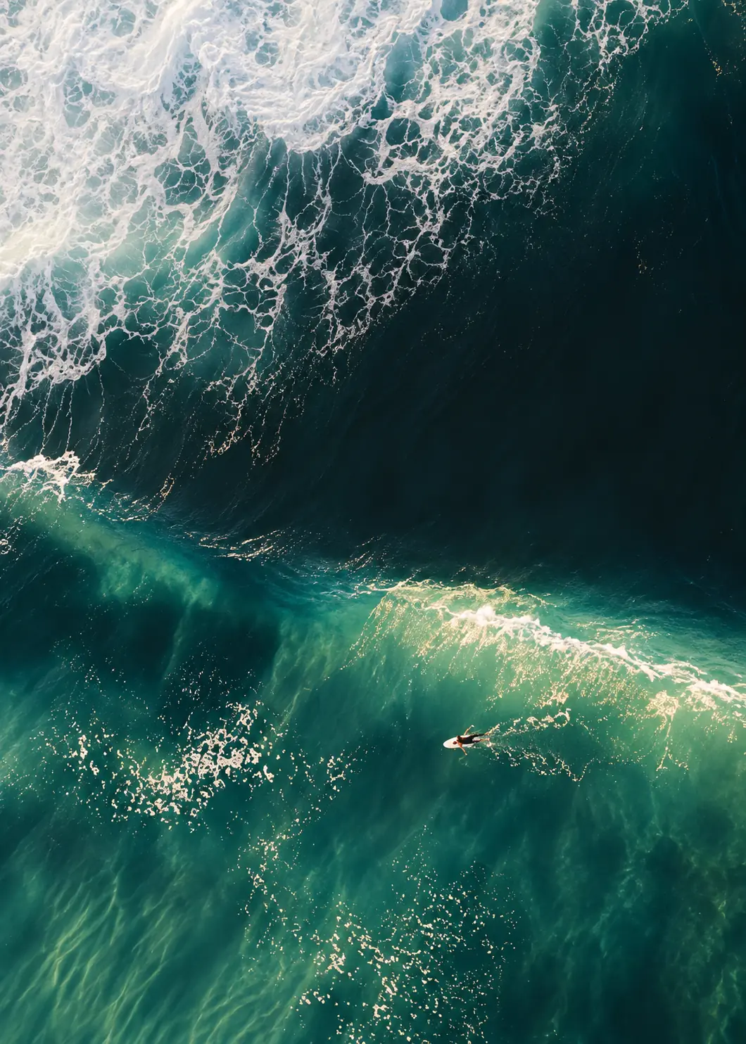 Aerial view of a deep green barrel wave with a tiny surfer and bright foam across the crest.