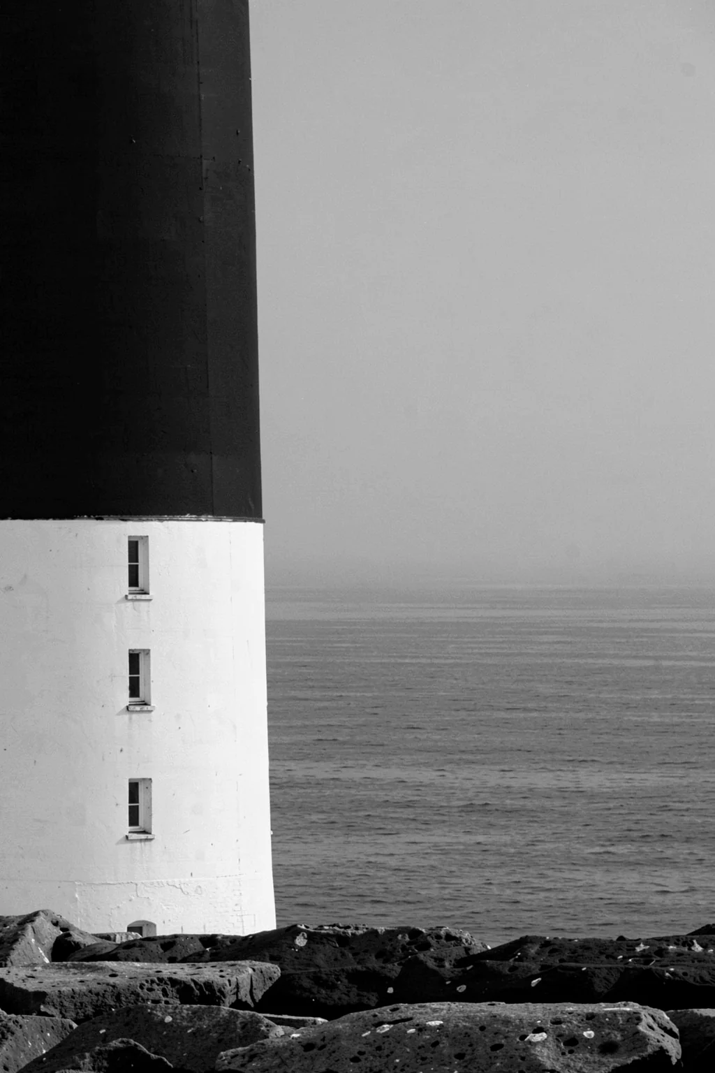 Beacon by the Sea. A cropped black-and-white lighthouse rises beside a calm ocean horizon above a rocky foreground.