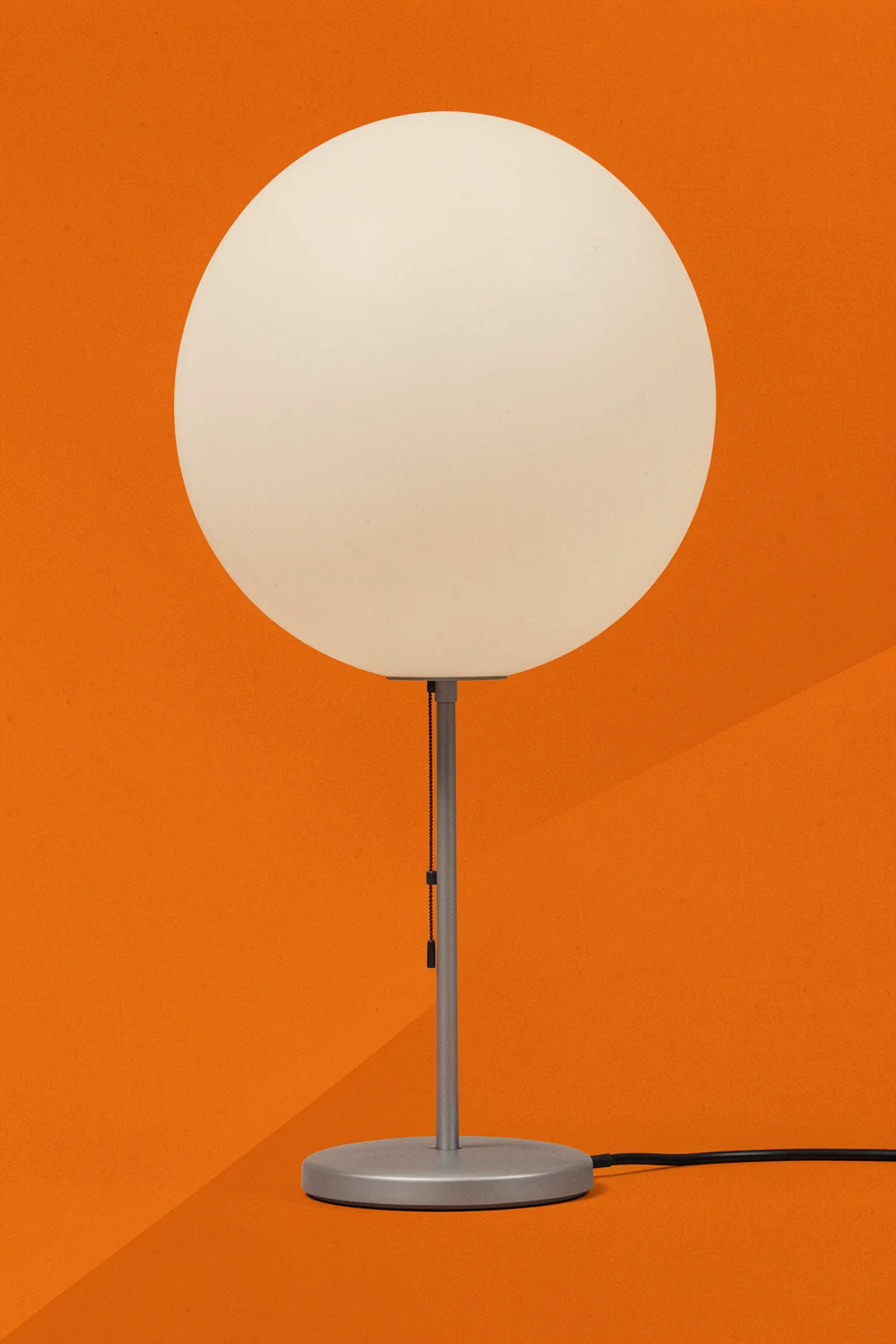 Wagenfeld Wg24. A slender silver table lamp with a large white globe shade is centered on a textured orange background.