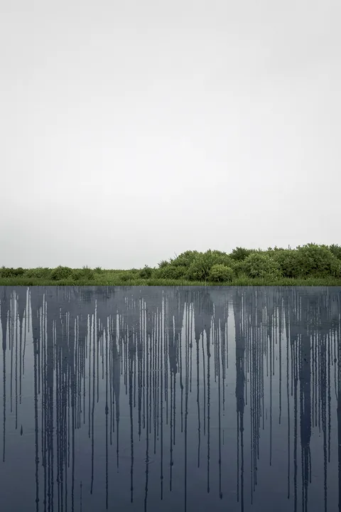 Serene Abstract Landscape. A narrow line of green shrubs borders dark blue water marked by long vertical drips, beneath a broad pale sky.