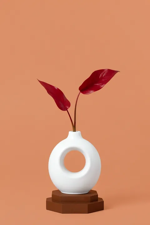 Abstract Botanical Vase. A white circular vase holding two dark-red leaves stands on a small brown plinth against a muted coral background.