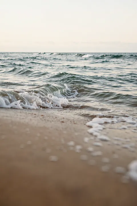 Abstract Waves. Small waves roll toward a sandy shoreline, with white foam in the foreground and a pale horizon beyond.