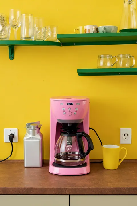 Back To Work. A pink drip coffee maker sits on a wooden counter beneath green shelves of cups and glassware, against a vivid yellow wall.