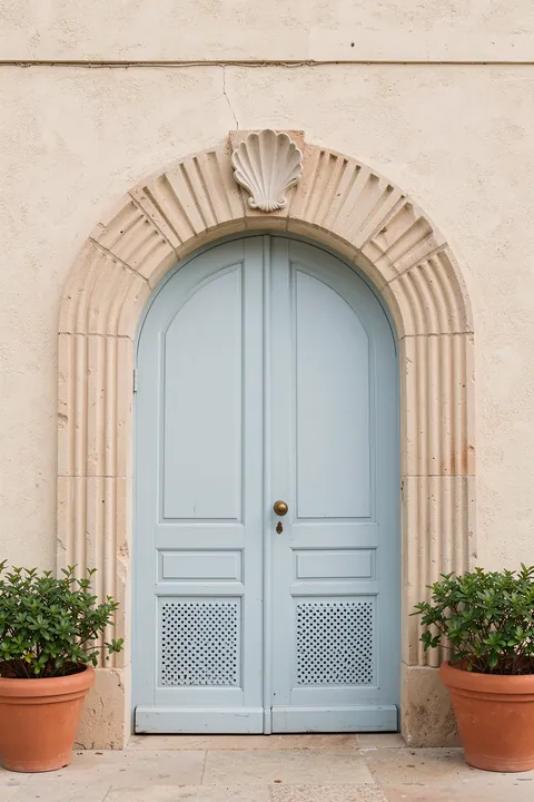 Blue tones of Rome - Italy travel photography. A pale stone arched doorway with a blue-gray door is framed by potted plants and greenery on a sunlit facade.