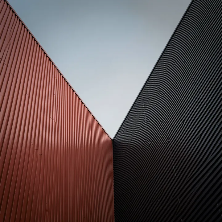 Dynamic Architectural Contrast. A narrow view between tall ribbed building facades contrasts rust red on the left with near-black on the right beneath a gray sky.