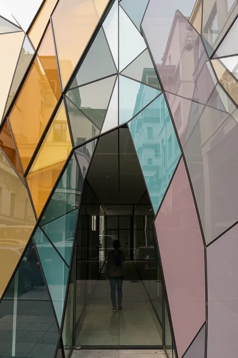 Geometric Glass Reflections. A person stands in a glass-fronted passage framed by intersecting angular window panes with muted colored reflections.
