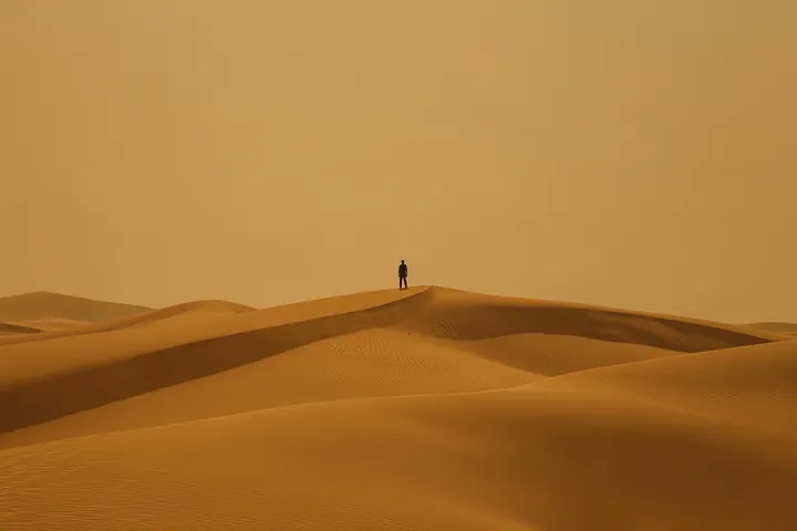 Golden Dune Solitude. A lone figure stands on a sunlit ridge above layered golden-orange dunes.
