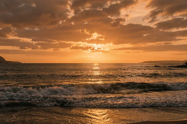 Golden Ocean Sunset. An orange sun sets over a dark ocean, with glowing light reflected across the water beneath clouded skies.