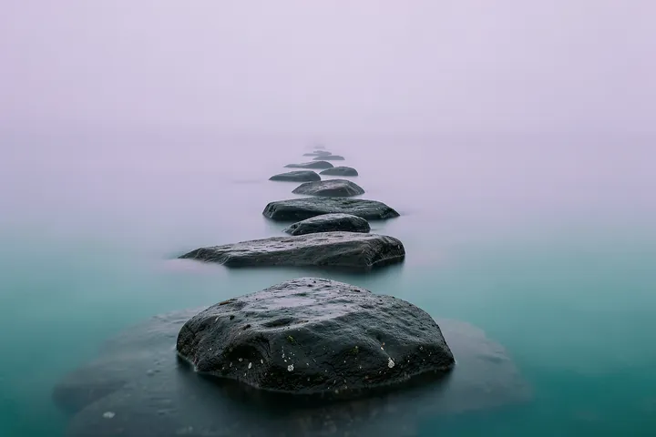 Misty Stone Path. A line of dark stepping stones recedes into smooth misty water beneath a pale lavender sky.