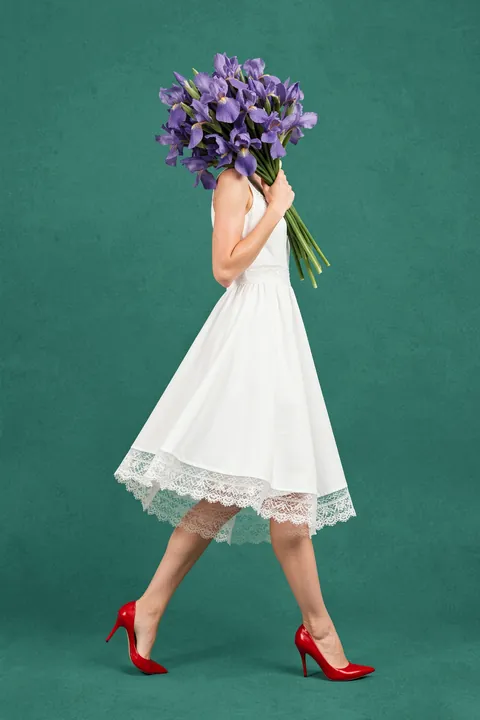 Stylish Floral Stroll. A woman in a white lace-trimmed dress walks across a deep teal field, holding a bouquet of purple flowers in front of her face; bright red heels add a vivid accent.