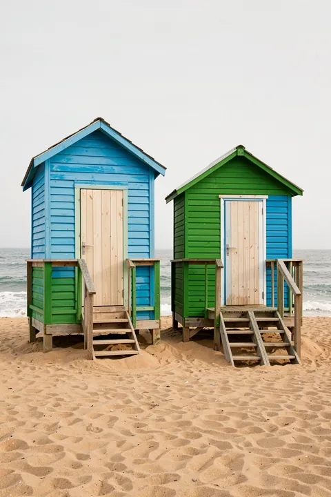 Coastal Huts Harmony. Two brightly painted beach huts stand on a sandy shore beside the sea, one blue and one green with blue side panels.