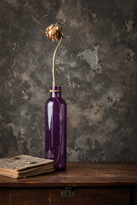 Wilted Bloom and Book. A dried flower in a purple glass bottle stands on a wooden surface beside an old book, against a mottled dark wall.