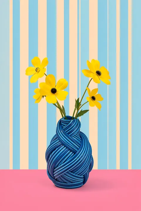 Yellow Flowers Knotted Vase. Yellow flowers rise from a sculptural, twisted dark blue vase, set against pale blue and cream stripes above a pink surface.