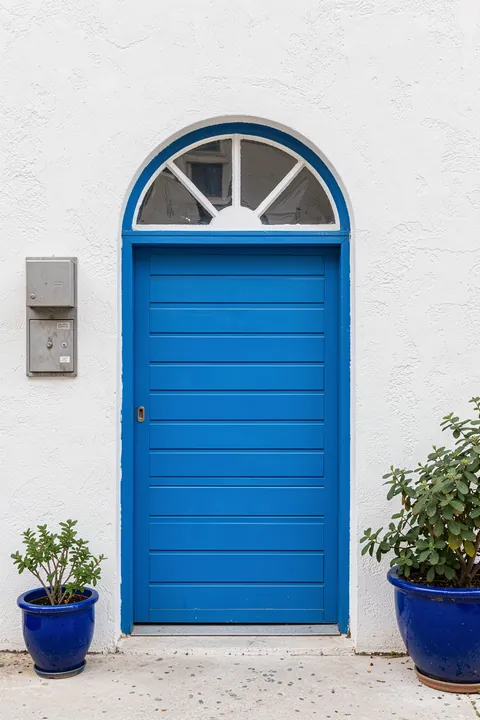Vibrant Blue Door. A bright blue paneled door is centered on a white stucco wall, flanked by leafy plants in blue pots.