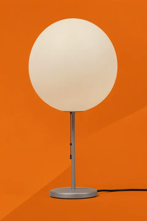Wagenfeld Wg24. A slender silver table lamp with a large white globe shade is centered on a textured orange background.