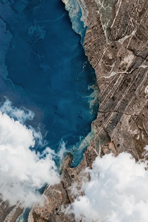 The Edge Upscaled Cgi 13287w. An aerial-style view shows deep blue water beside a rugged brown shoreline, partly veiled by white clouds.
