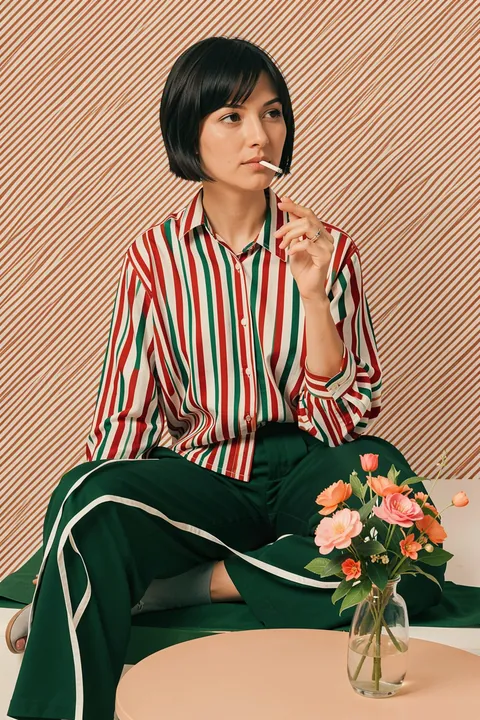 Woman in Stripes Shirt and Green Pants. A seated woman in a red, green and cream striped shirt and dark green trousers is shown beside a small vase of flowers.