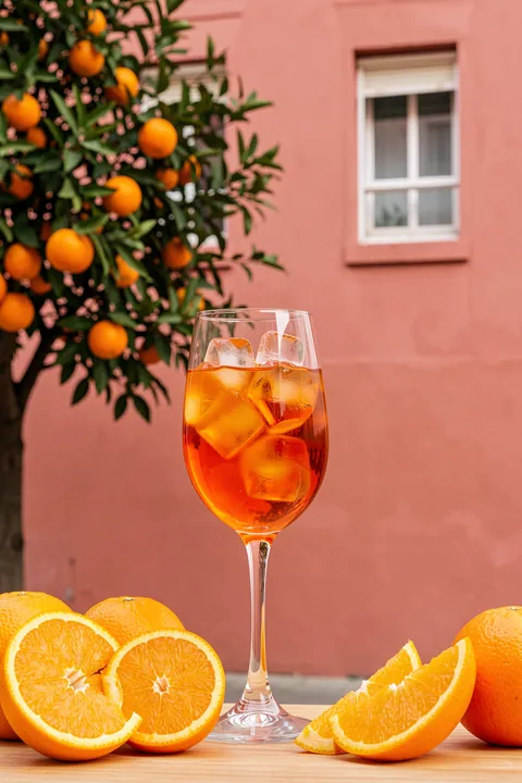 Drink Summer Scene. A glass of iced amber drink is surrounded by orange slices and fruit-bearing branches against a pink wall.