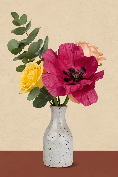 Arranged Flowers. A vivid magenta poppy, yellow rose and pale peach blooms are arranged with eucalyptus in a speckled ceramic vase.
