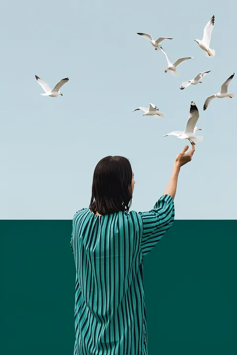 Woman Embracing Freedom. Seen from behind, a woman in a teal striped dress reaches toward a flock of seabirds above dark teal water.