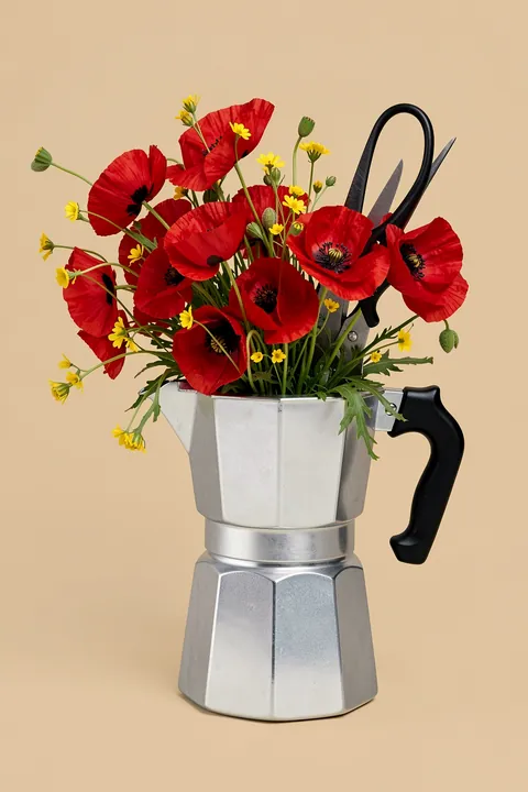 Coffee Garden Blooms. Bright red poppies and small yellow flowers are arranged in a silver moka pot with a black handle against beige.