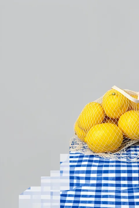 Fresh Citrus Still Life. A mesh bag of bright yellow lemons rests on blue-and-white checked cloth against a pale grey wall.