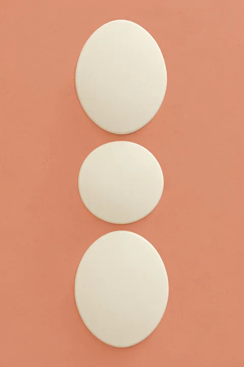 Earthy Stacked Vessels. Three raised ivory oval forms are stacked vertically on a muted coral background.
