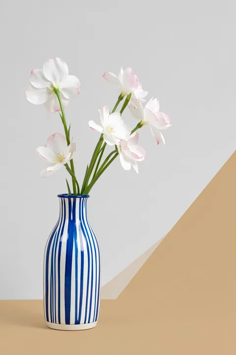 Blue Striped Flower Vase. A slim blue-and-white striped vase holds delicate white flowers beside simple tan geometric shapes.