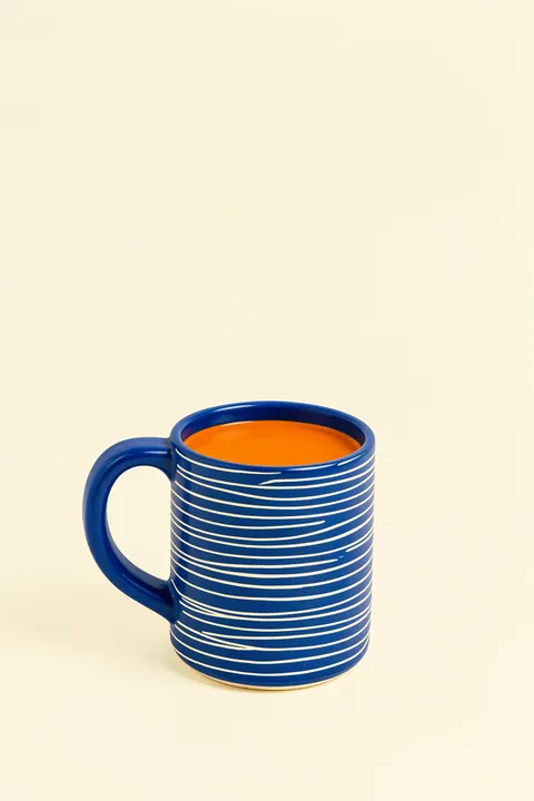Blue Mug Moment. A blue mug with thin white stripes, filled with orange liquid, sits against a pale cream background.