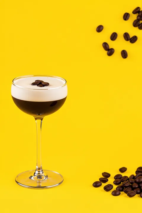 Bold Espresso Cocktail. A dark espresso martini with a creamy foam top and coffee beans is served in a stemmed glass against bright yellow.