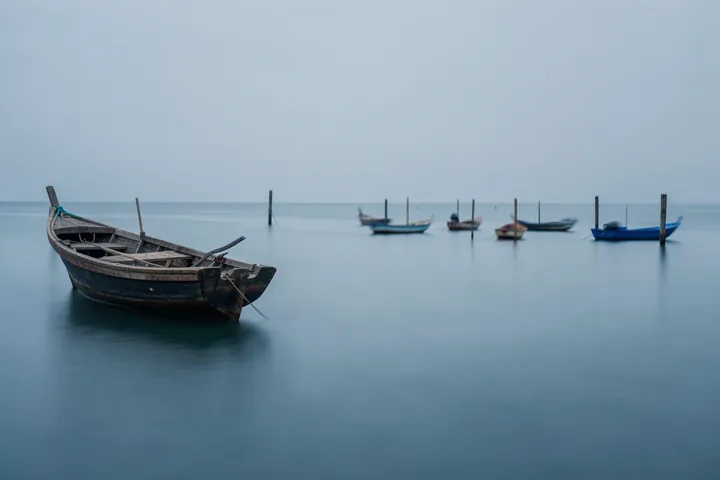 Calm Water Boats. A weathered wooden boat rests on still blue-gray water, with smaller boats fading into the distant horizon.