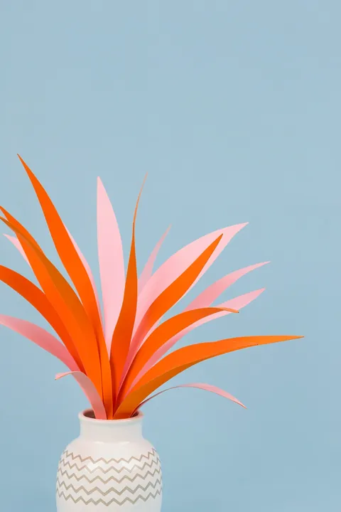 Minimal Botanical Still Life. Long orange and pink leaves fan from a white patterned vase against a pale blue background.