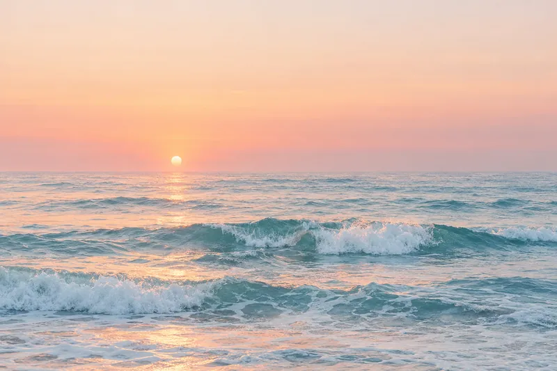 Pink sunrise over a turquoise sea with small white waves breaking on a sandy beach.